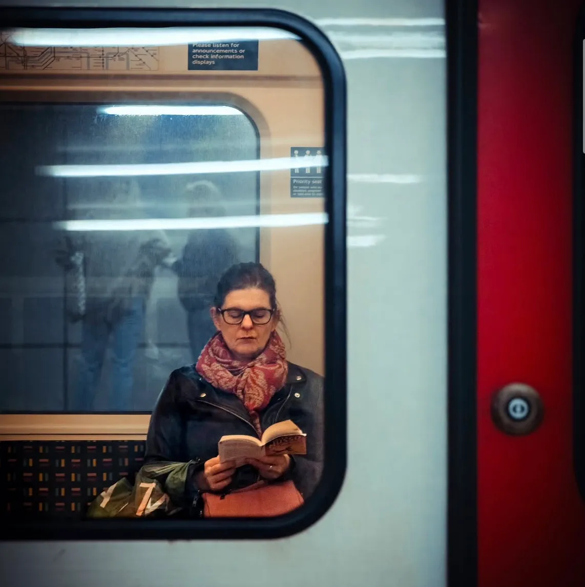 A woman reading a paperback, framed by the window of a stationary Central line carriage, reflections of the platform sliding across the glass.