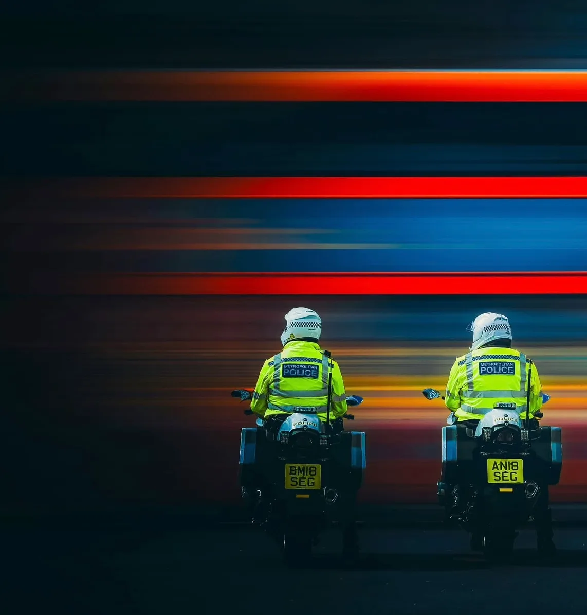 Two police motorcyclists in high-visibility jackets held still against a wall of red and blue light trails on a London street at night.