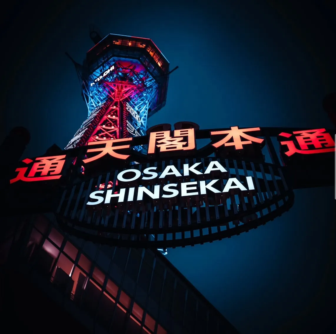 The Tsūtenkaku tower seen from directly beneath, framed by the illuminated red Shinsekai archway against a deep blue night sky.